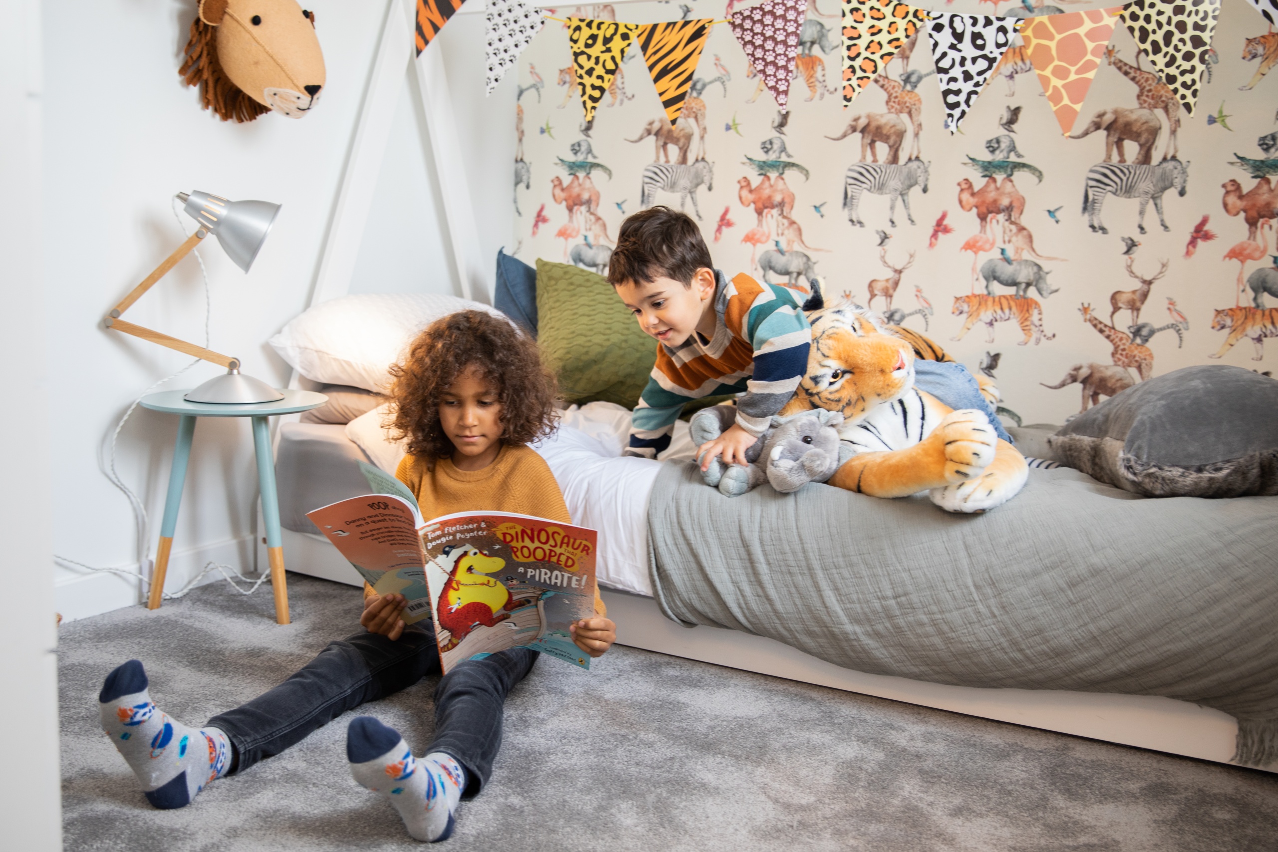 Two children sat with each other in a bedroom reading a children's book about dinosaurs